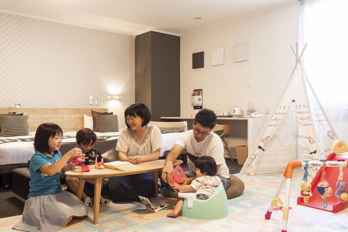 Una familia en una habitación de hotel con cama, escritorio, silla, zona de juegos con juguetes y ventana con cortinas.