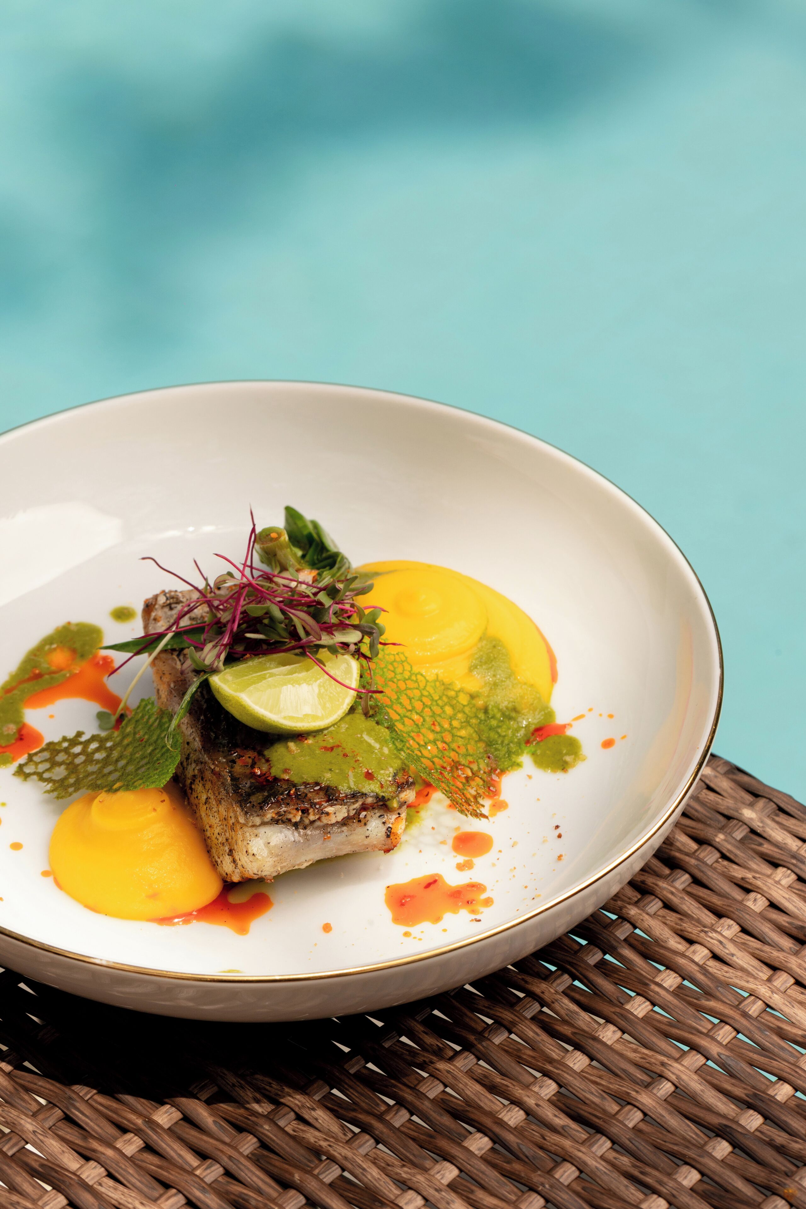 A white plate with a fish dish, garnished with greens and a lime slice, on a woven mat.
