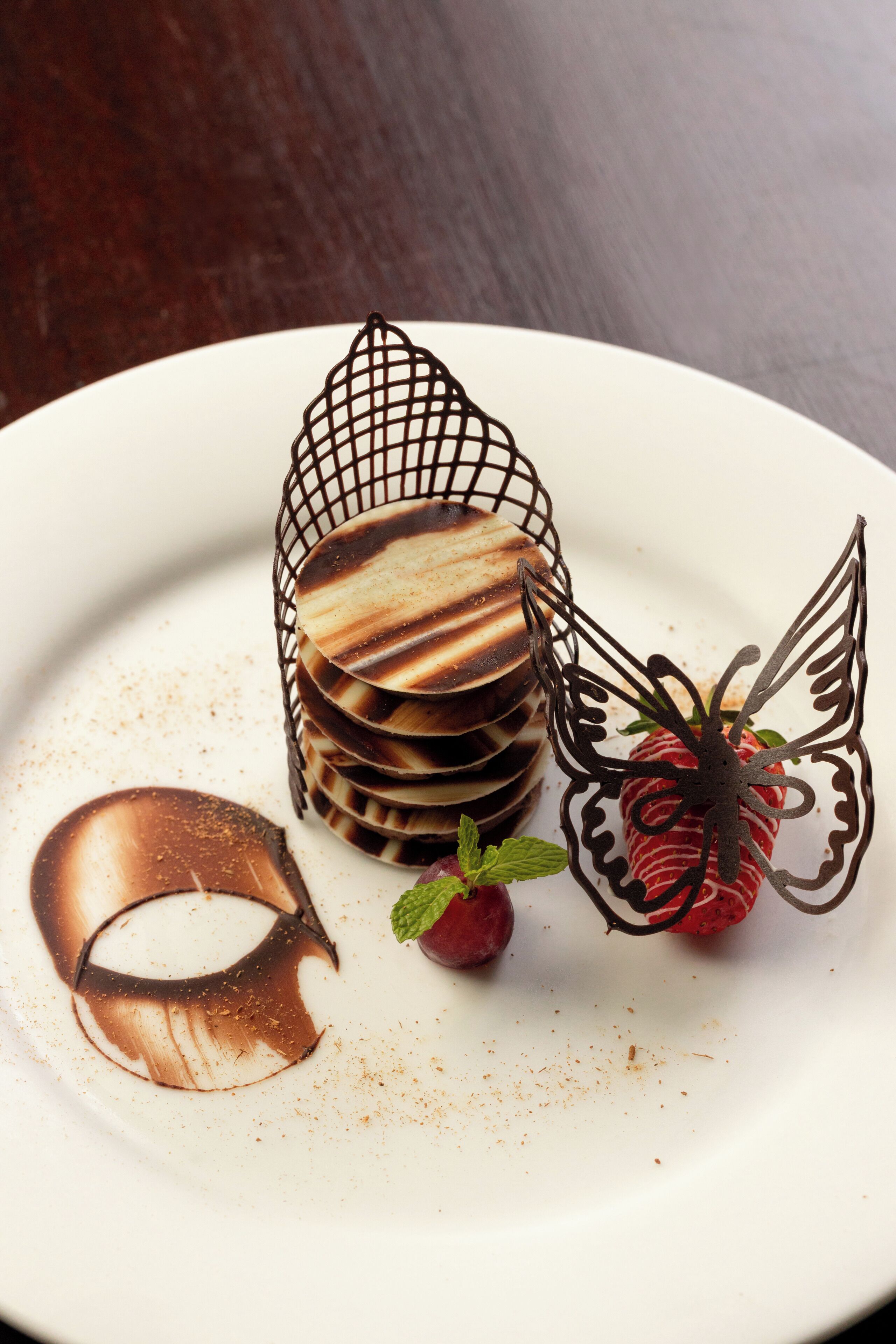 Une assiette à dessert ornée d’un motif en chocolat, une pile de gaufrettes au chocolat et une fraise décorée d’un papillon en chocolat.