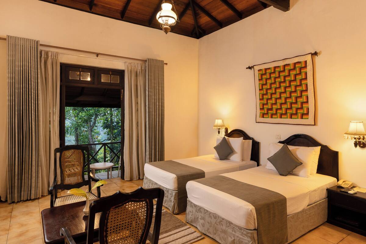 A hotel room with two beds, a chair, a table, a window with curtains, and a patterned wall hanging.