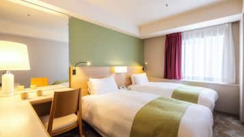 A hotel room with two beds, a desk with a lamp, a chair, and a large window with curtains.
