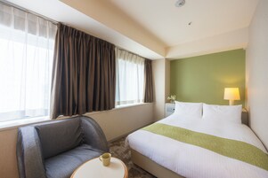 A hotel room with a bed, a chair, a table with a cup, and a lamp.