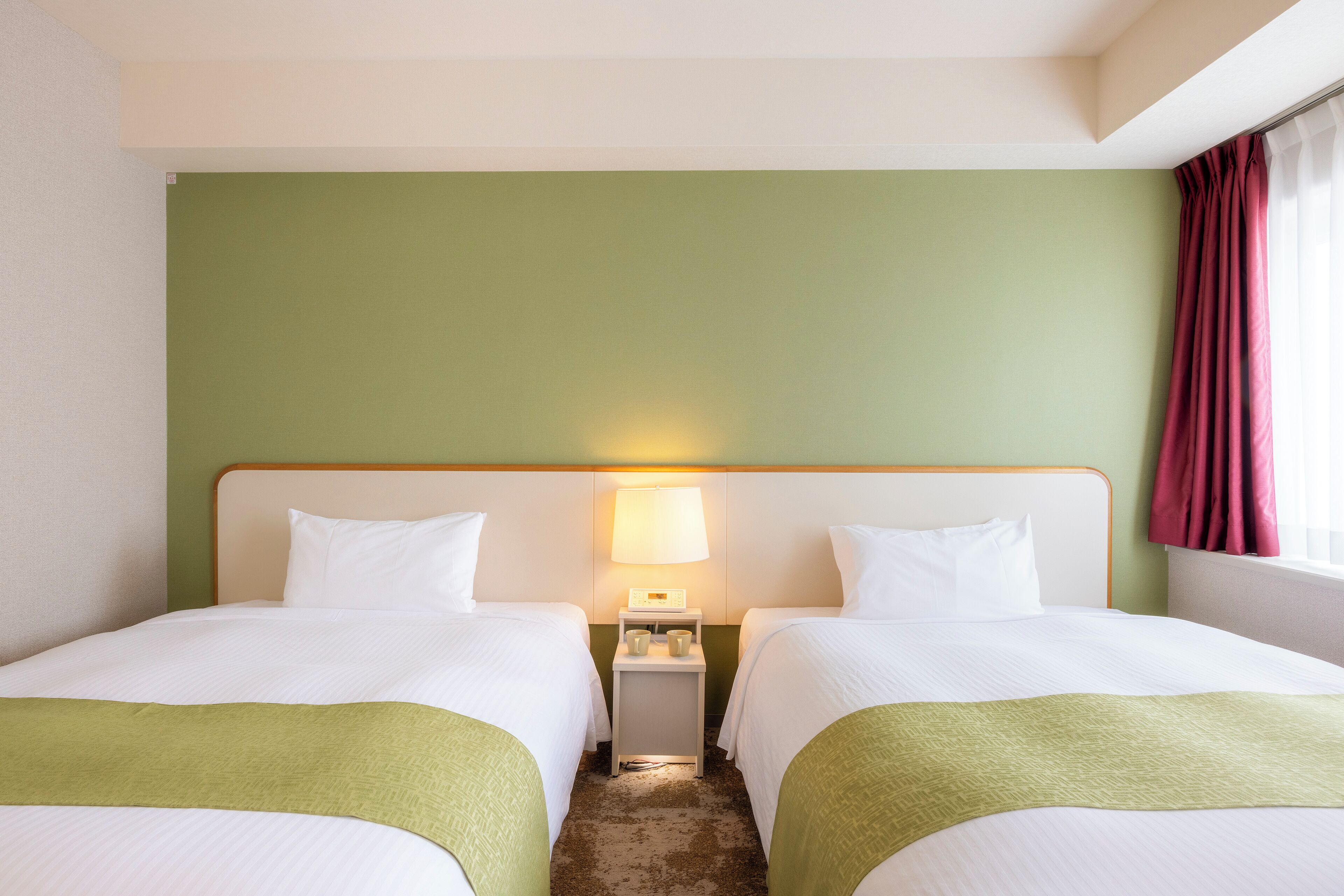 A hotel room with two beds, a nightstand with a lamp, and a window with curtains.