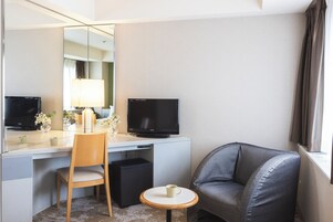 A hotel room with a desk, two monitors, a chair, a small table with a cup, and a sofa.