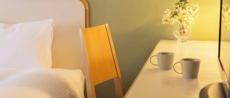 Habitación de hotel con una cama, una silla de madera, una mesa con dos tazas y un florero con flores.