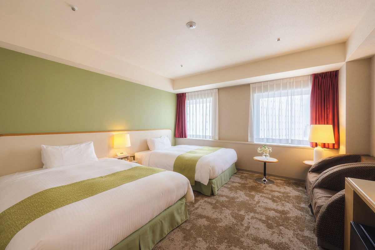 Ein Hotelzimmer mit zwei Betten, einem kleinen Tisch, einem Stuhl und einem Fenster mit Vorhängen.