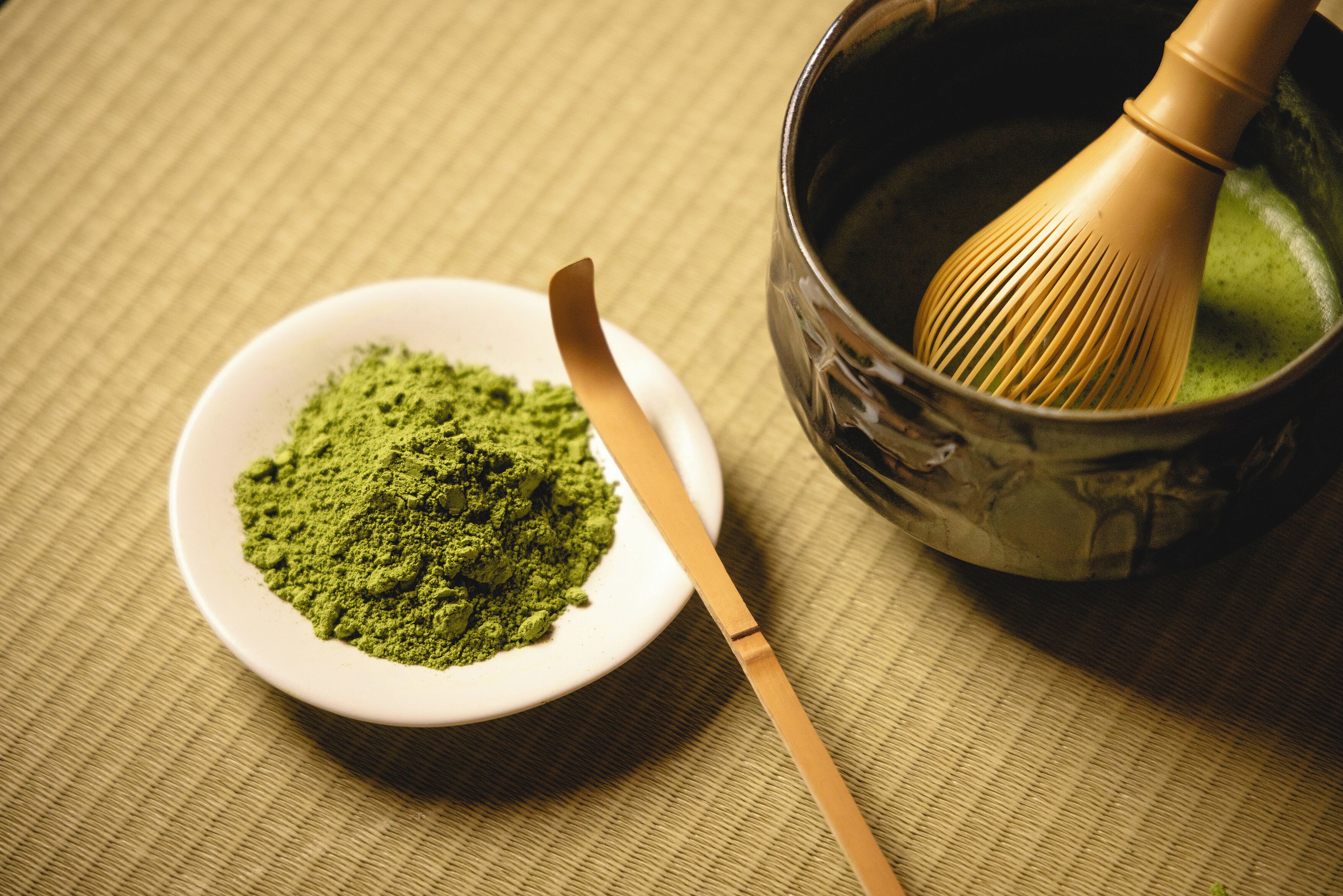 A bowl of green powder, a whisk, and a bowl of liquid on a textured surface.