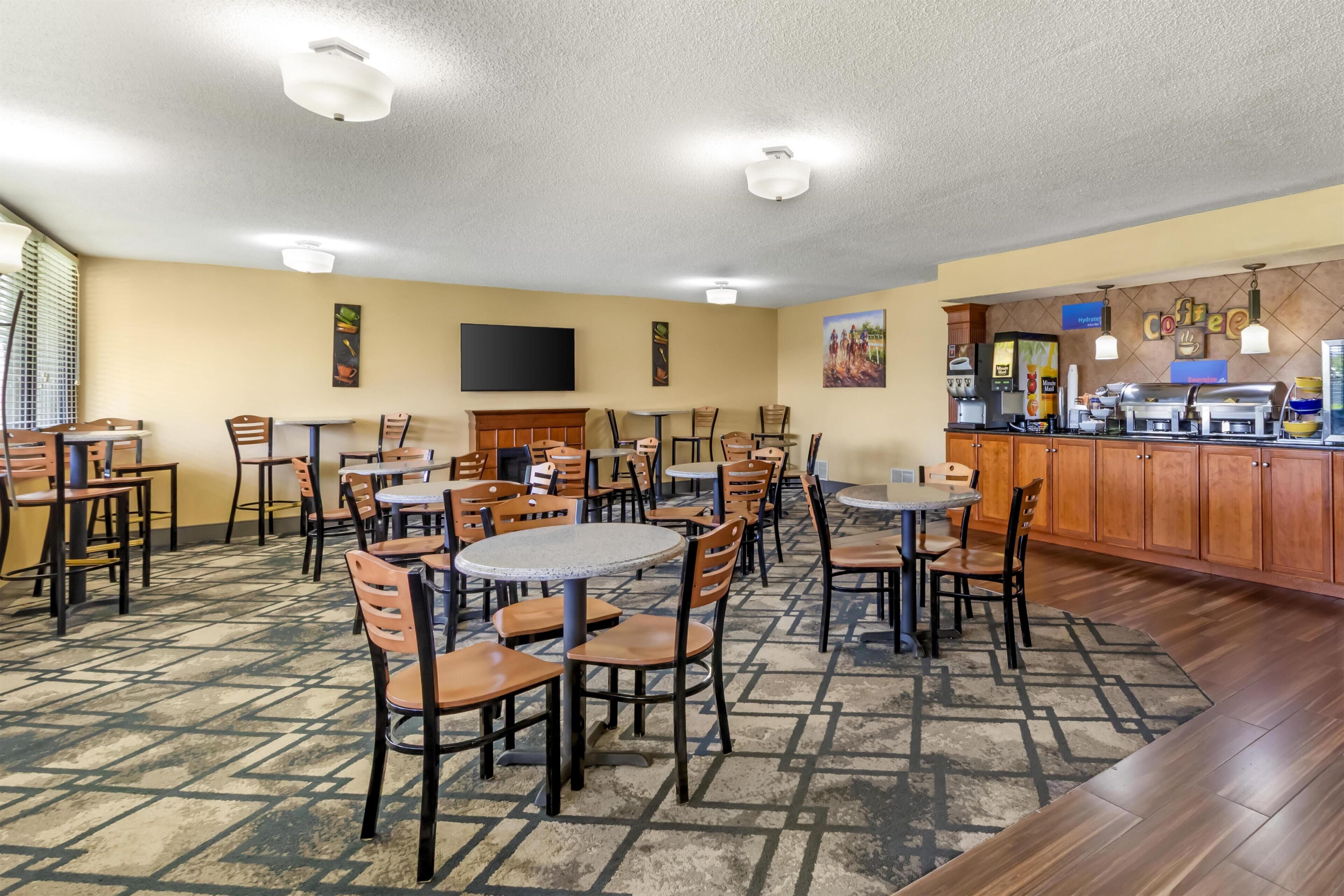A hotel dining area with multiple tables and chairs, a coffee station, and a flat-screen TV on the wall.