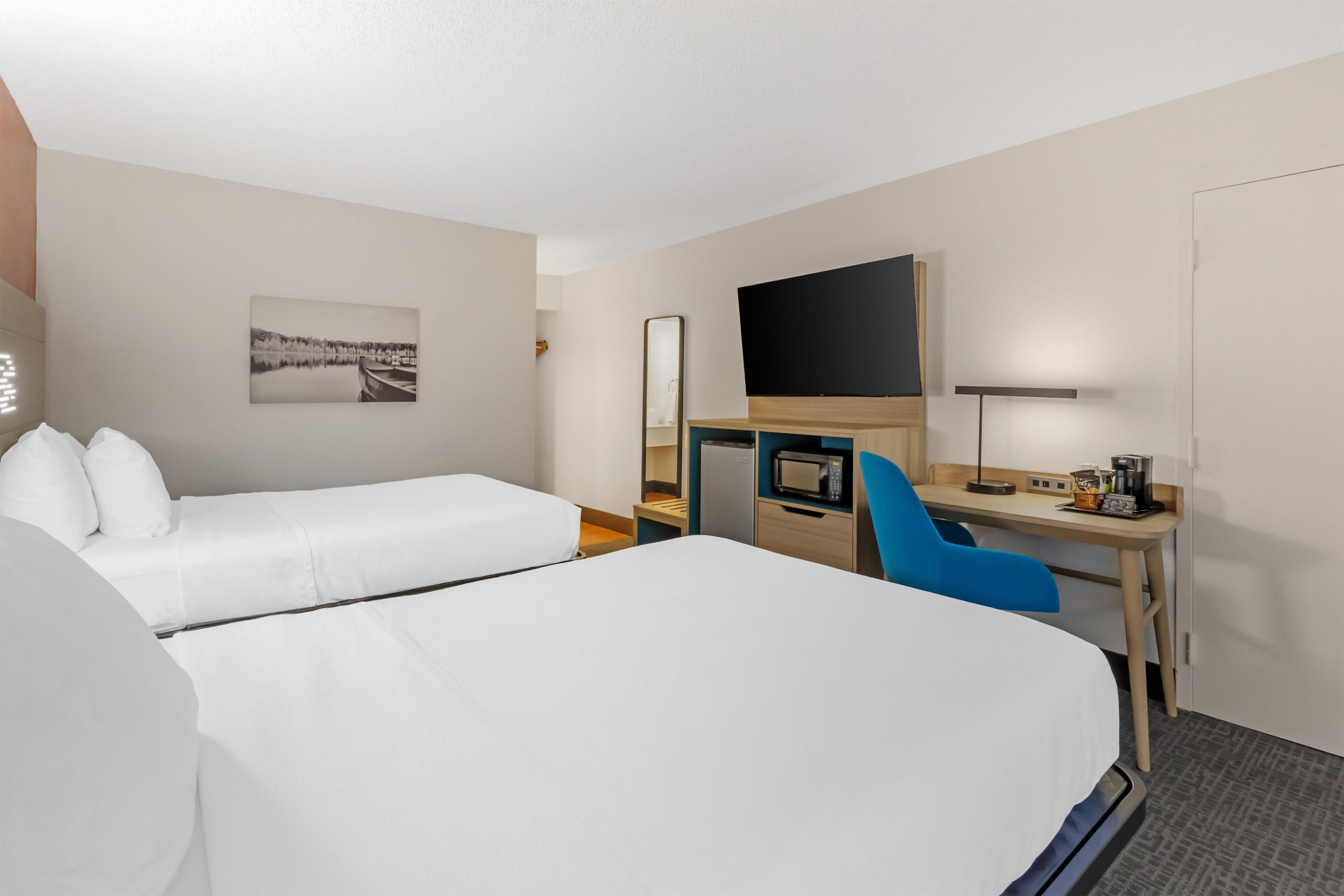 Habitación de hotel con dos camas, un escritorio, una silla, un televisor de pantalla plana, un espejo y un cuadro en la pared.