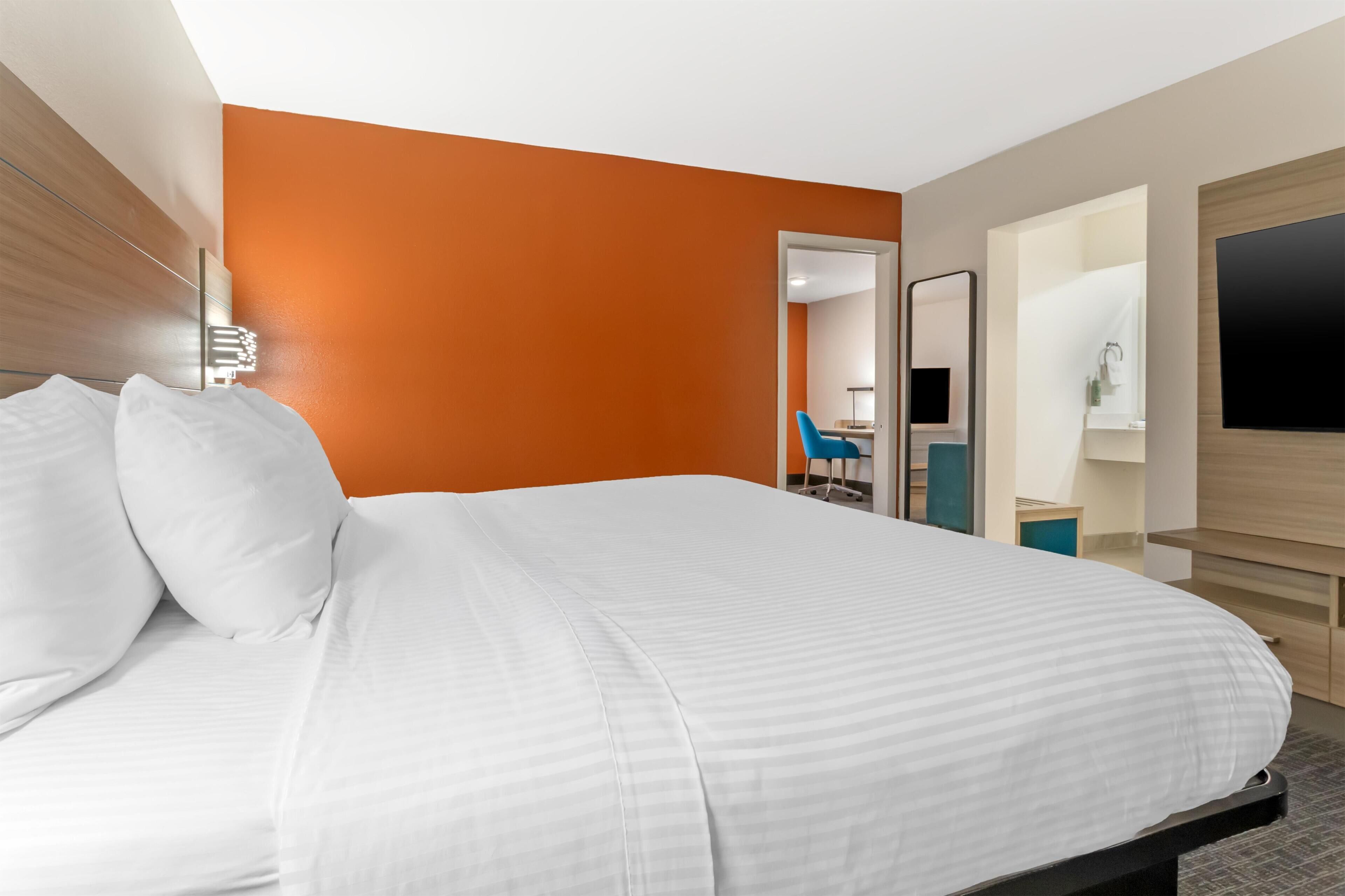 Habitación de hotel con cama, espejo, televisor y baño visible a través de una puerta abierta.