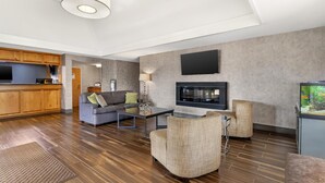 A modern living room with a sofa, armchairs, a coffee table, a flat-screen TV, and a kitchen area with wooden cabinets.