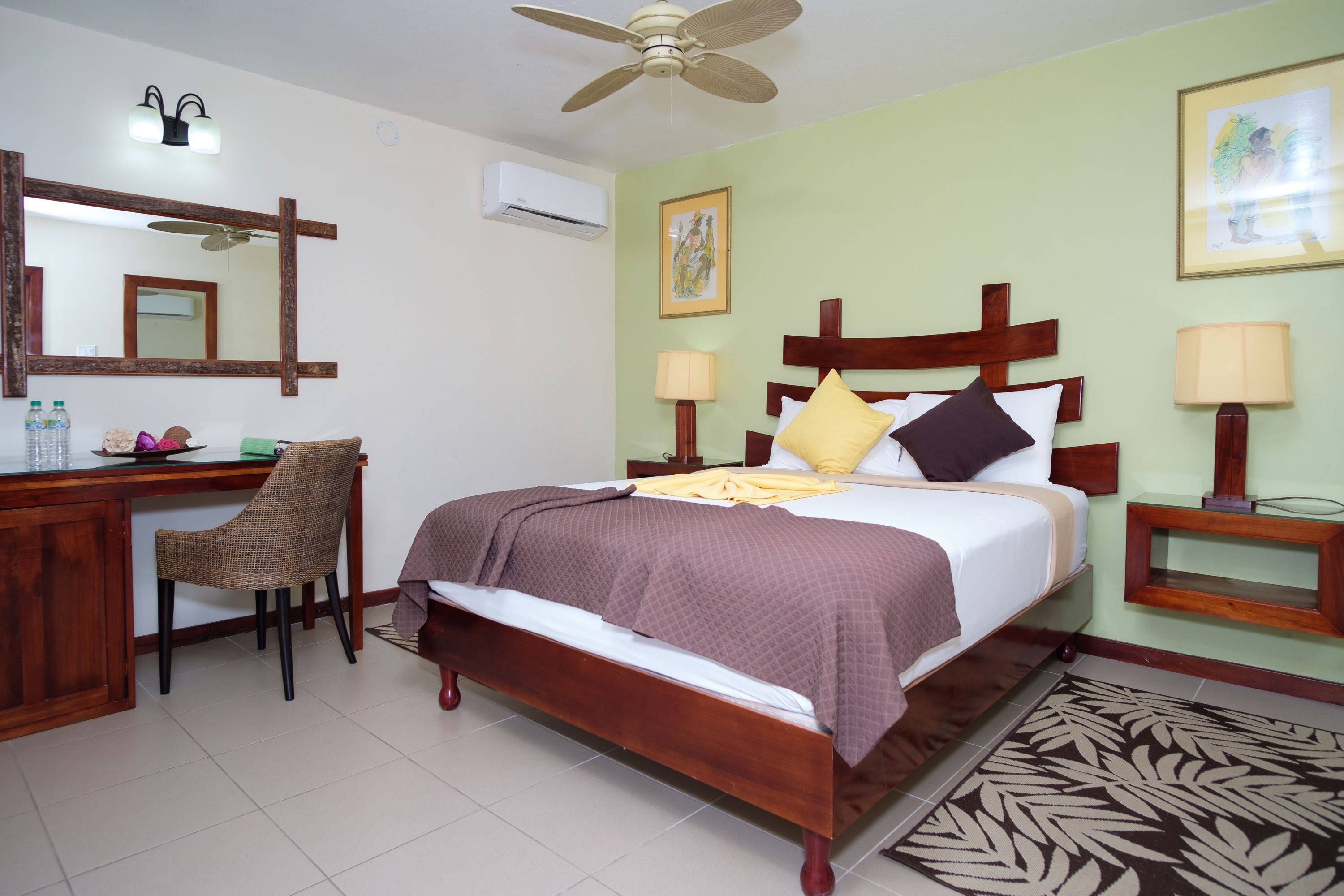 Una habitación de hotel con una cama de madera, un escritorio con una silla, un ventilador de techo y un espejo grande.