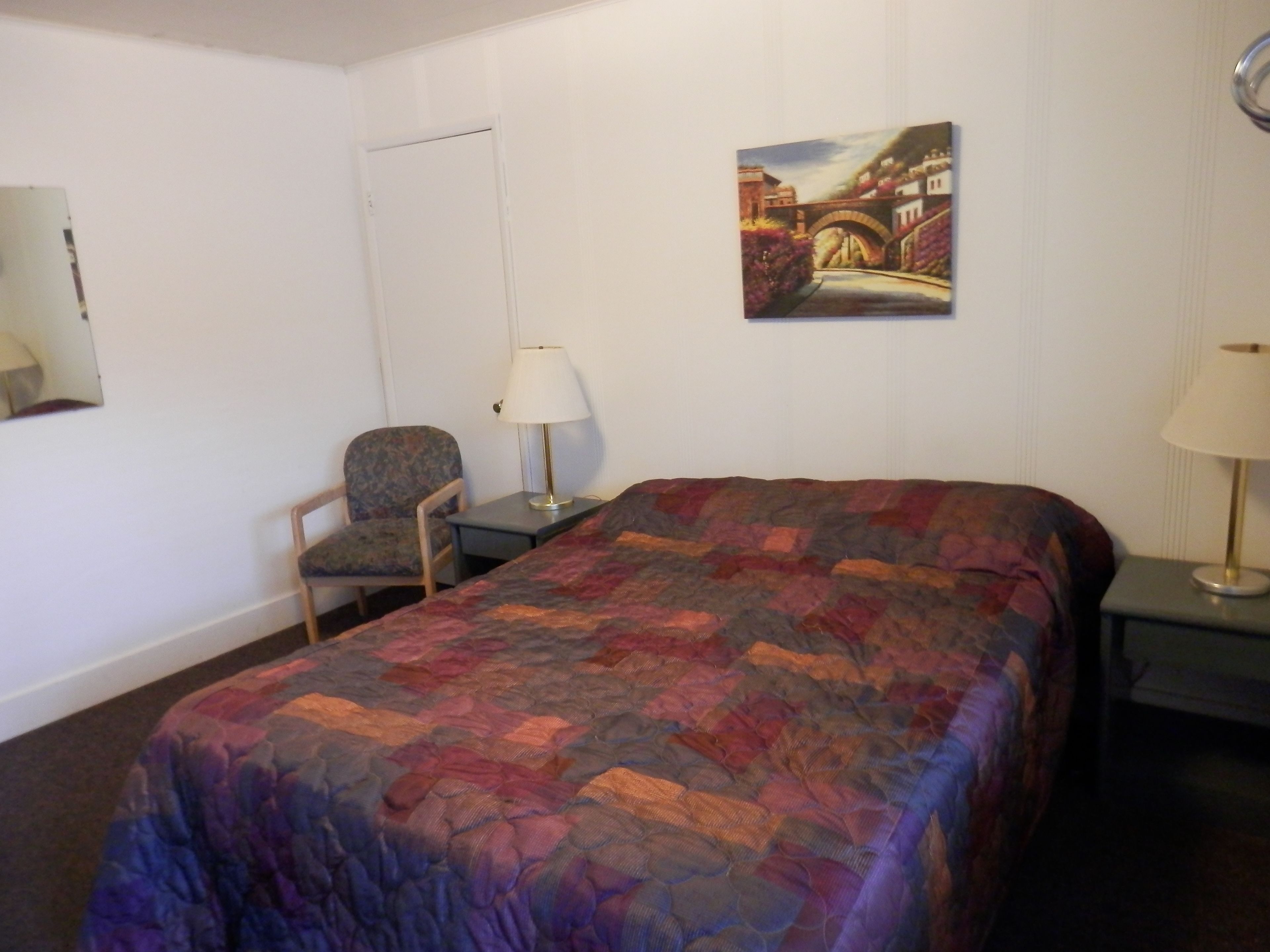 A hotel room with a bed, a chair, a desk, a lamp, and a painting on the wall.