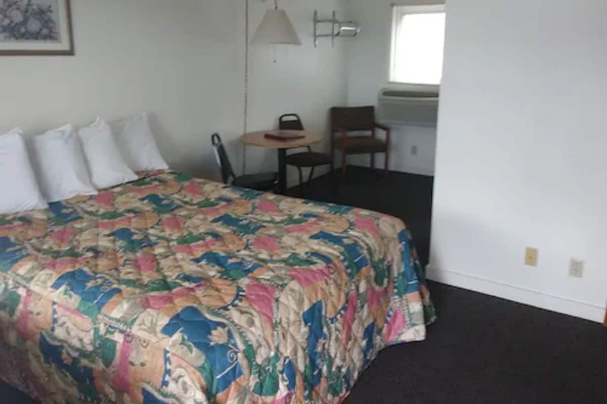 Quarto com cama, almofadas, escrivaninha, cadeira, luminária e janela.
