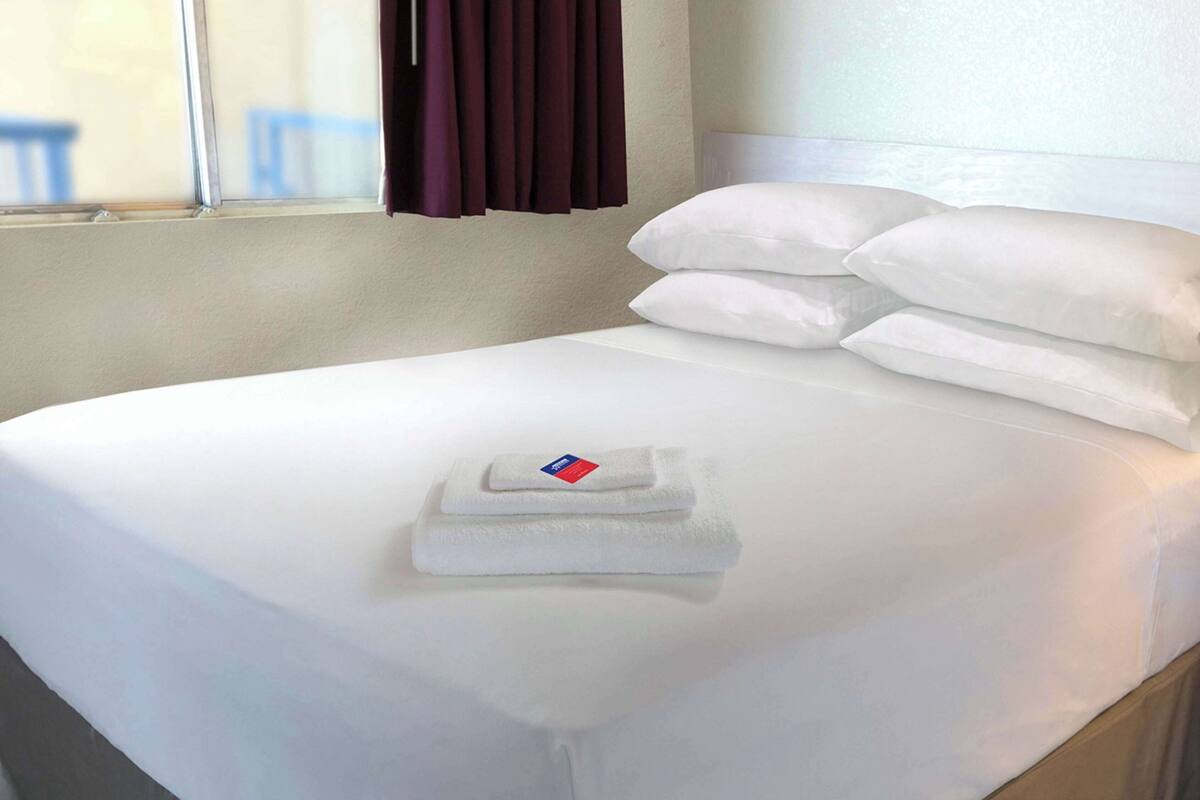 A neatly made bed with white linens and pillows, a folded towel on the bed, and a window with curtains.