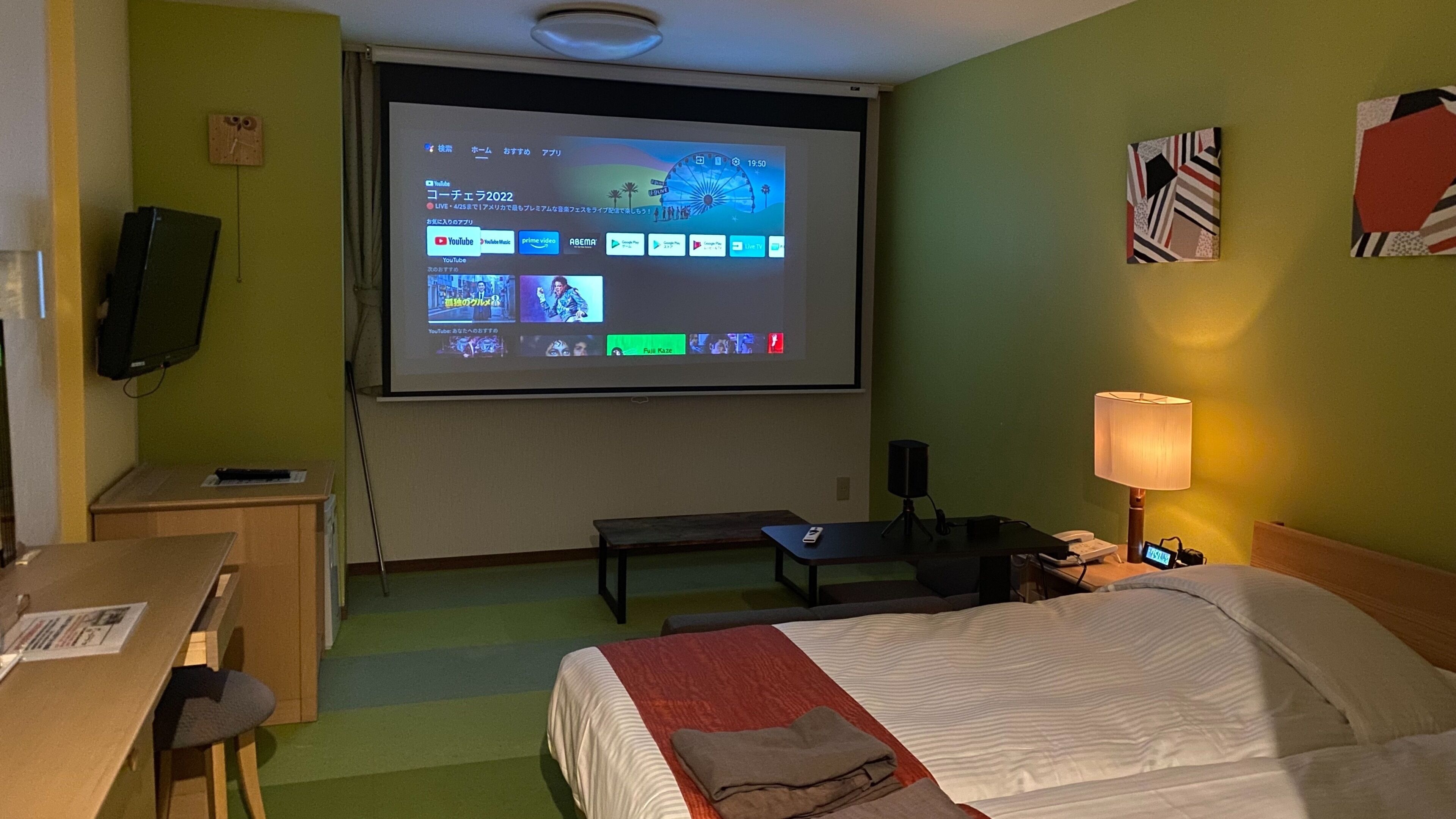 Habitación de hotel con cama, escritorio, silla, televisor mostrando un video, lámpara y reloj en la pared.
