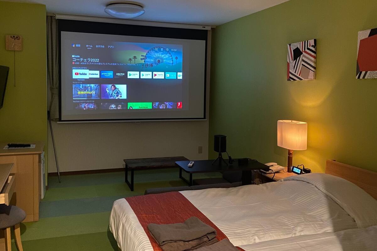A hotel room with a bed, a desk, a chair, a TV screen displaying a video, a lamp, and a clock on the wall.