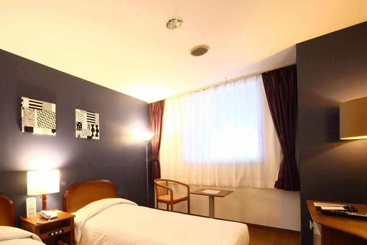 Une chambre d’hôtel avec deux lits, un bureau, une chaise, une lampe et une fenêtre avec des rideaux.
