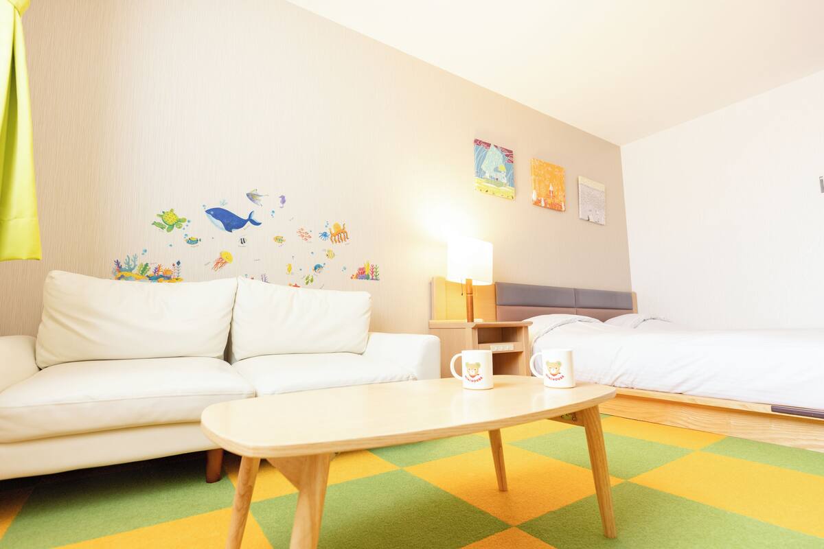 A hotel room with a white sofa, a small table with two mugs, a lamp, and a bed.