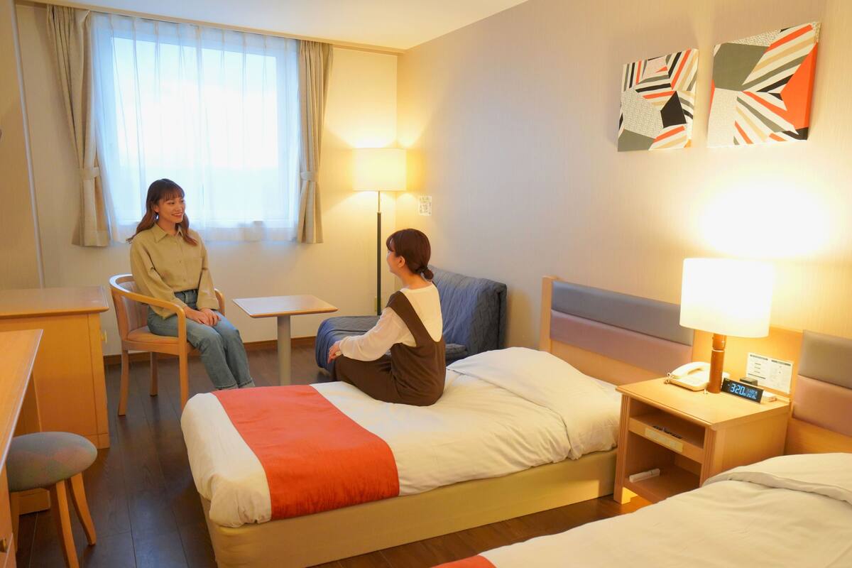 A hotel room with a bed, a desk, a chair, and a nightstand with a lamp.