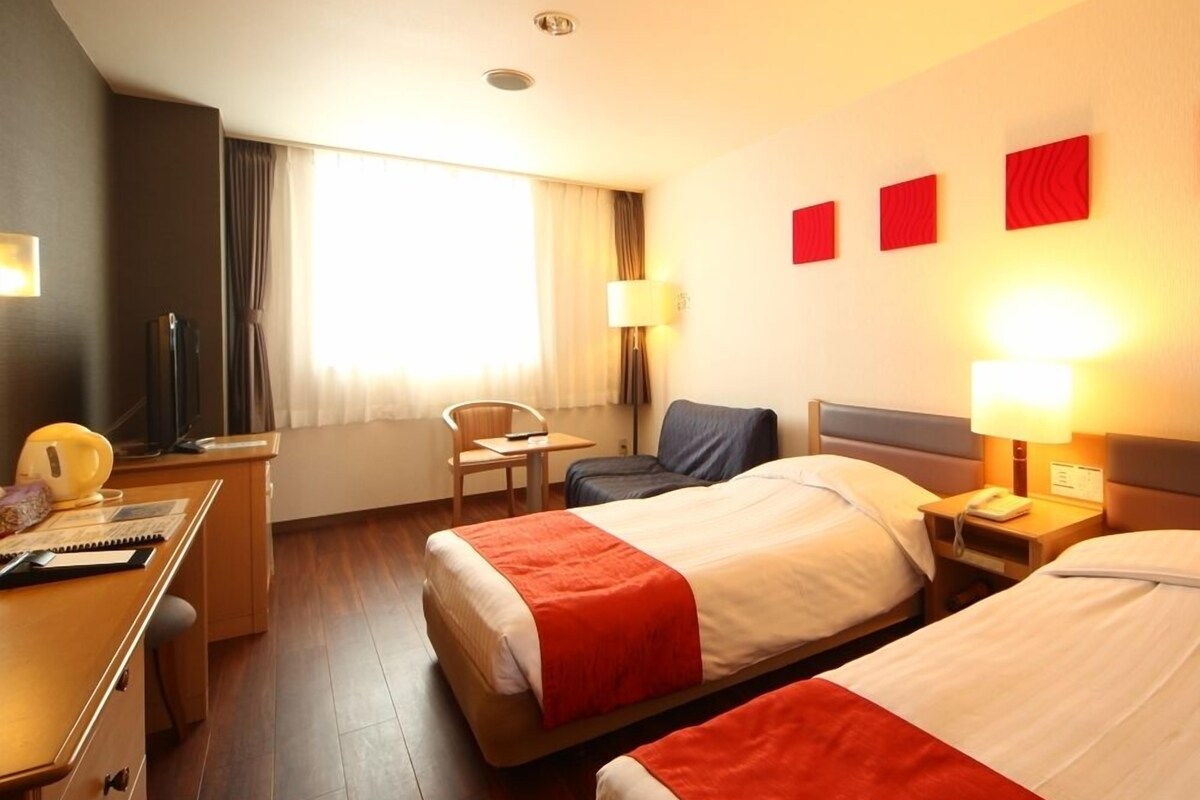 Une chambre d’hôtel avec deux lits, un bureau, une chaise, une lampe et une fenêtre avec des rideaux.