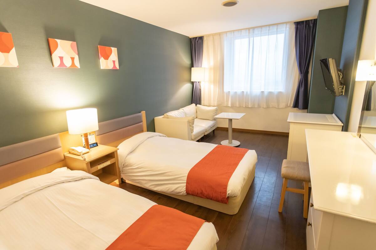 A hotel room with two beds, a desk, a chair, a small table, and a lamp.