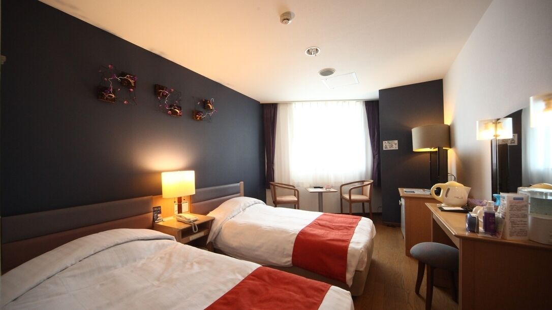 A hotel room with two beds, a desk, a chair, a lamp, and a window with curtains.
