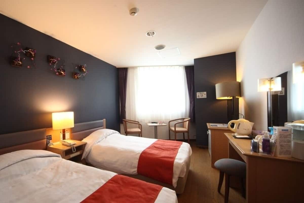 A hotel room with two beds, a desk, a chair, a lamp, and a window with curtains.