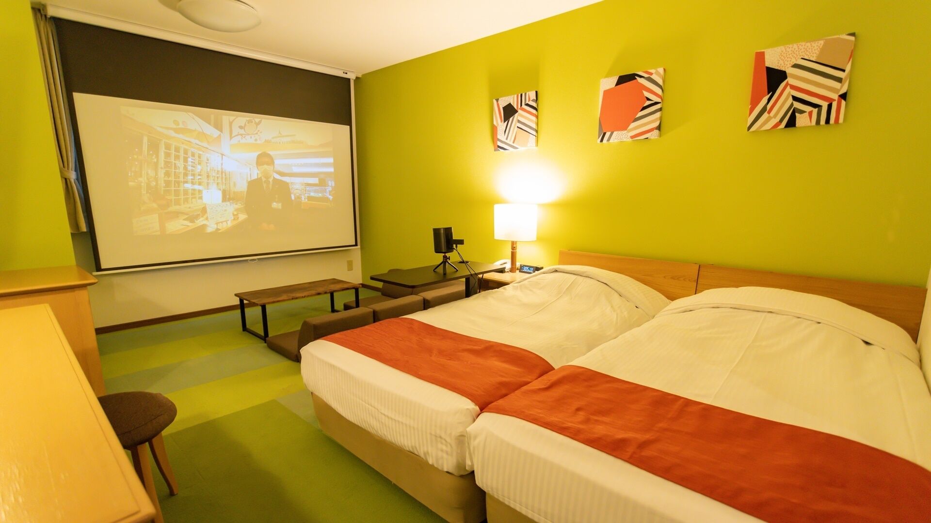 A hotel room with a bed, a desk, a projector screen, and a lamp.