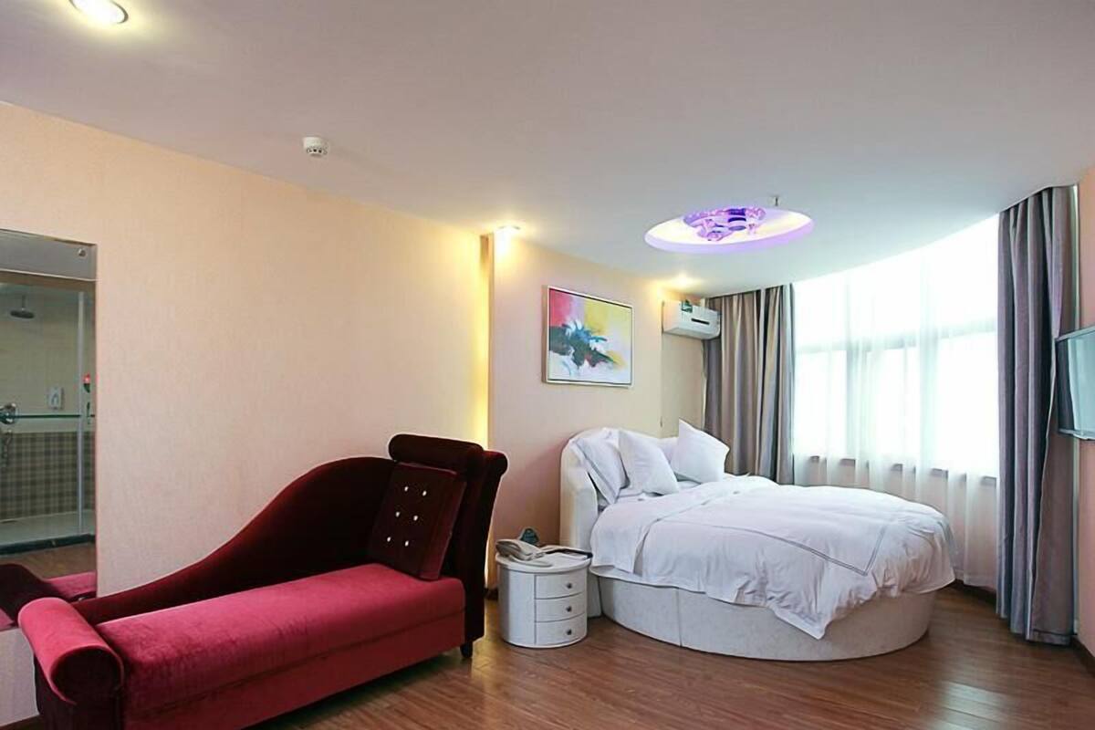 Ett hotellrum med en säng, en röd soffa, ett litet sängbord, en tv som är monterad på väggen och ett fönster med gardiner.