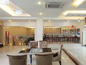 A hotel lobby with a central dining area, a staircase, and a reception desk.