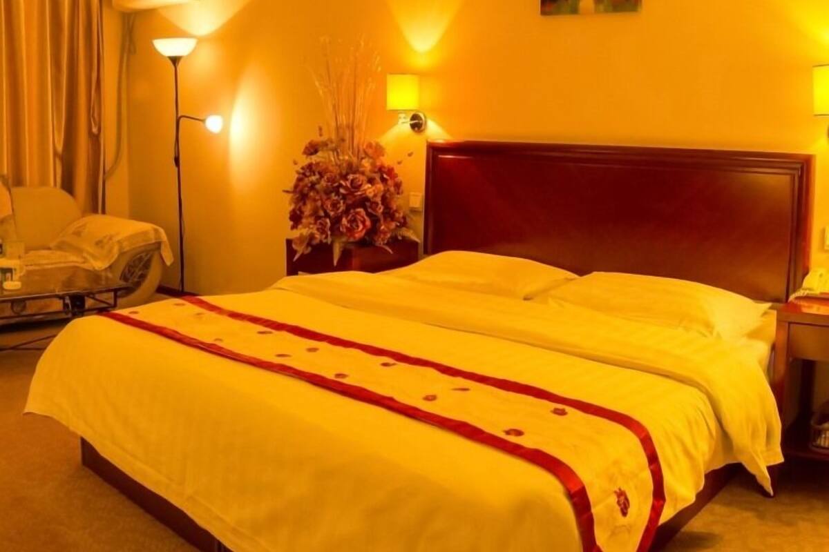 Una habitació d'hotel amb un llit gran, una taula de nit amb una làmpada, una cadira i un vas amb flors.