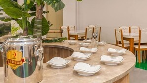 Un comedor con una mesa redonda dispuesta para cuatro personas, rodeada de sillas de madera. Un gran macetero está en primer plano, y una jarra de metal decorativa sobre la mesa.
