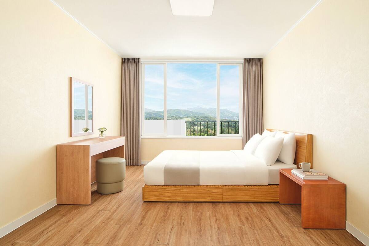 Una habitación de hotel moderna con piso de madera, una cama con ropa blanca, un escritorio con una silla y un ventanal con vista a las montañas.