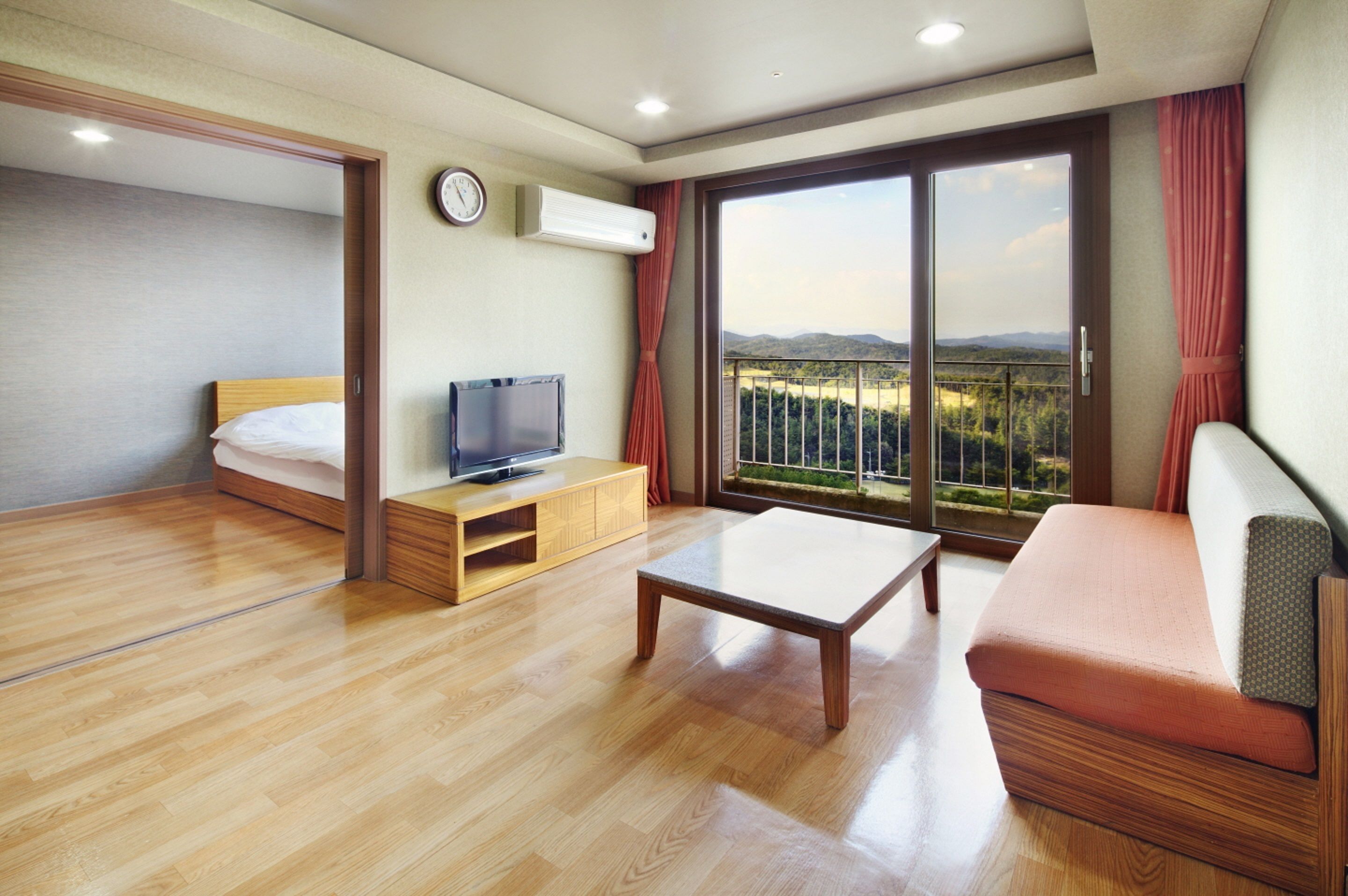 A hotel room with a bed, a TV, a sofa, a wooden coffee table, and a large window with a view of the outdoors.