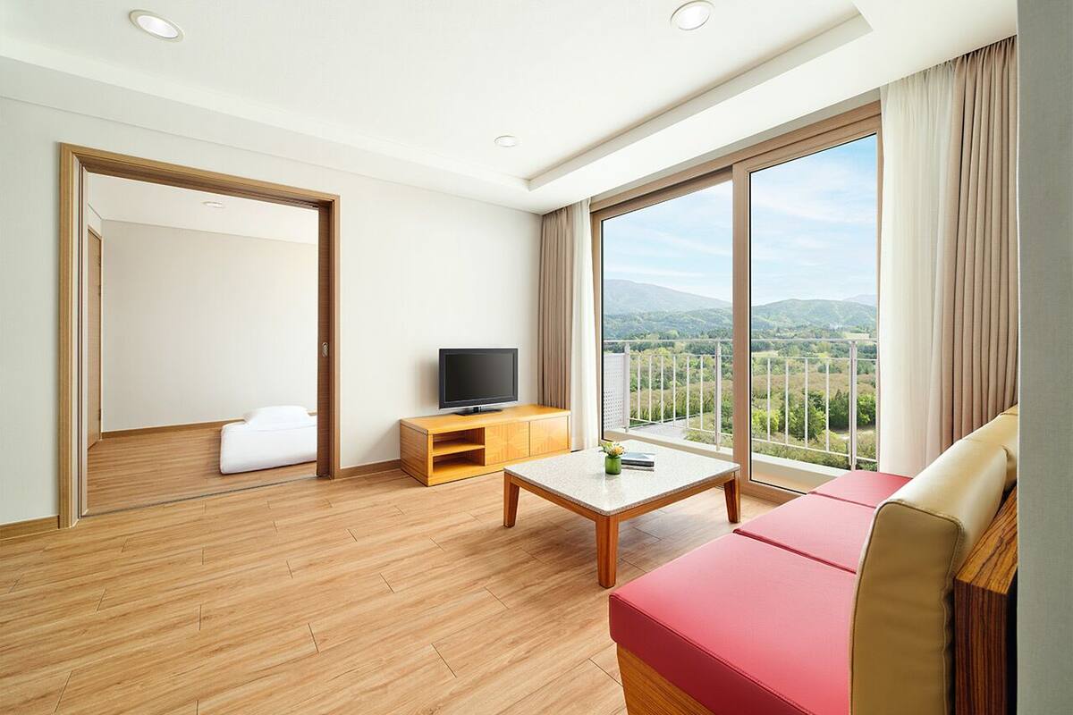 Una habitación de hotel moderna con suelo de madera, un televisor de pantalla plana sobre un soporte de madera, un sofá rojo y un ventanal con vistas al campo.