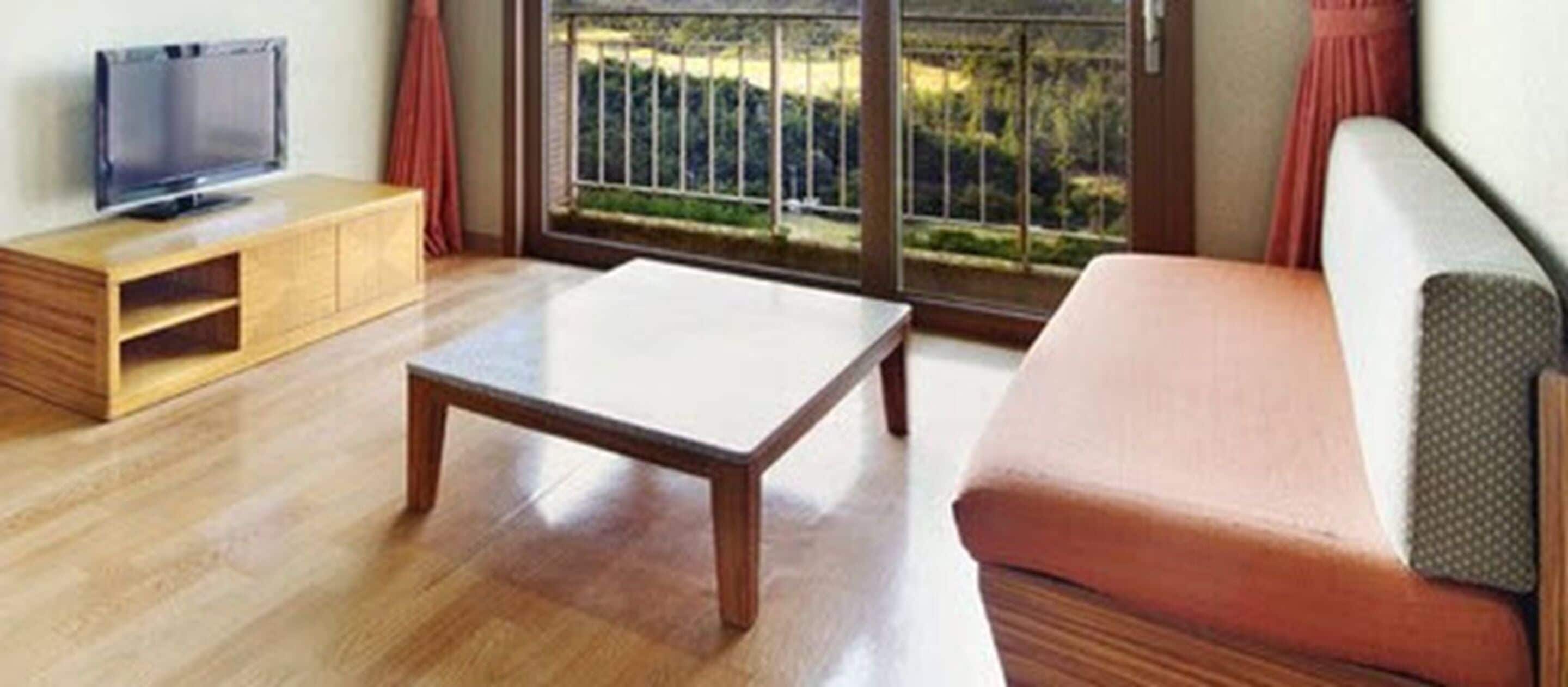 A living room with a wooden coffee table, a sofa, a TV on a wooden stand, and a large window with a view of greenery.