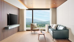 Un salon moderne avec un téléviseur à écran plat fixé sur un mur recouvert de panneaux en bois, une table basse en bois, un fauteuil beige et un canapé gris offrant une vue sur les montagnes grâce à de grandes fenêtres.