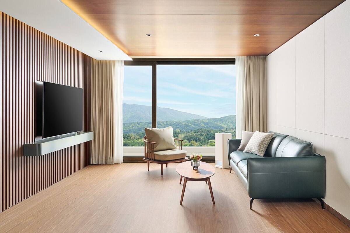 Una sala de estar moderna con un televisor de pantalla plana montado en una pared de paneles de madera, una mesa de centro de madera, un sillón color beige y un sofá gris con vistas a montañas a través de grandes ventanales.