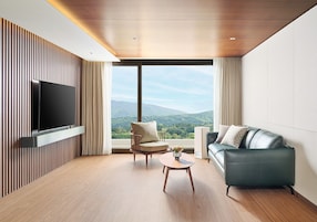 Una sala de estar moderna con un televisor de pantalla plana montado en una pared de paneles de madera, una mesa de centro de madera, un sillón color beige y un sofá gris con vistas a montañas a través de grandes ventanales.