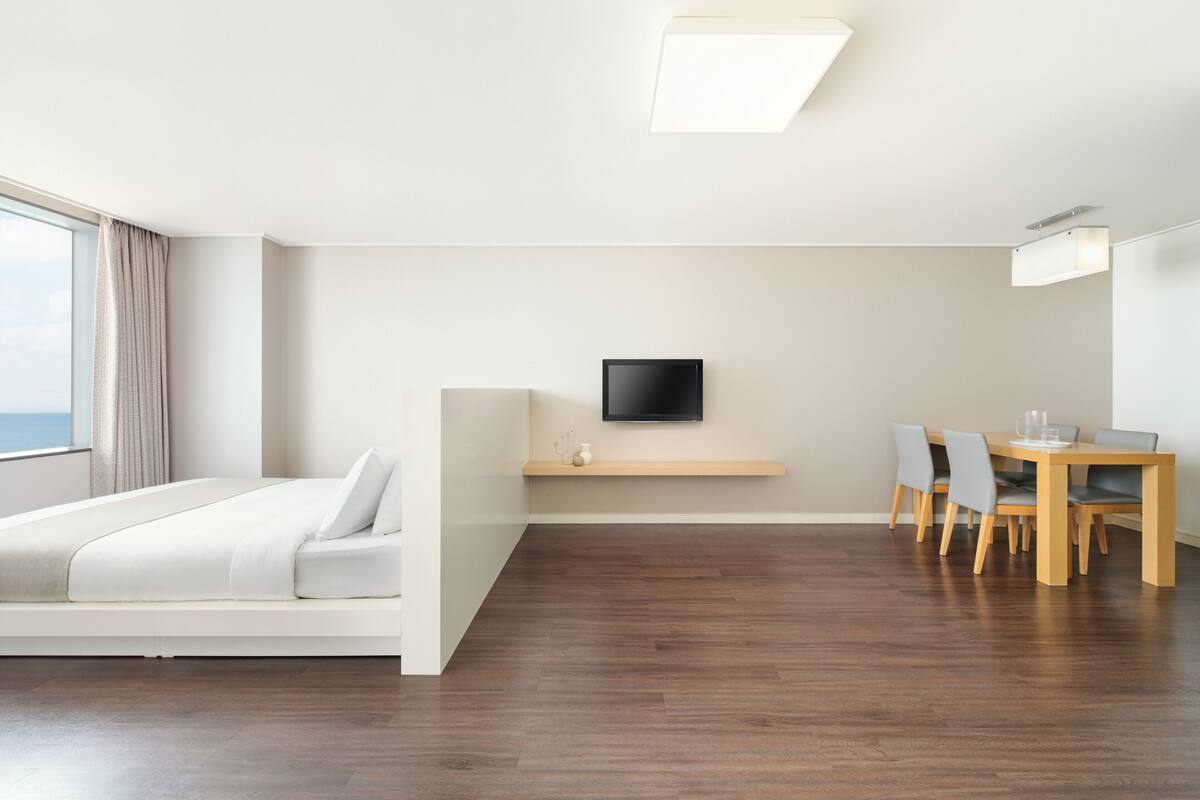 Een moderne hotelkamer met een bed, een aan de muur bevestigde televisie, een eettafel met stoelen en uitzicht op zee.