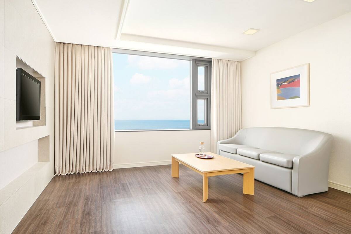 Een moderne woonkamer met een houten vloer, een beige bank, een klein houten tafeltje, een aan de muur bevestigde televisie en een groot raam met gordijnen.