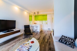 Una sala de estar moderna con televisor de pantalla plana, una mesa de centro de vidrio con revistas, un sofá con un cojín decorativo y una cocina con armarios verdes y electrodomésticos blancos.
