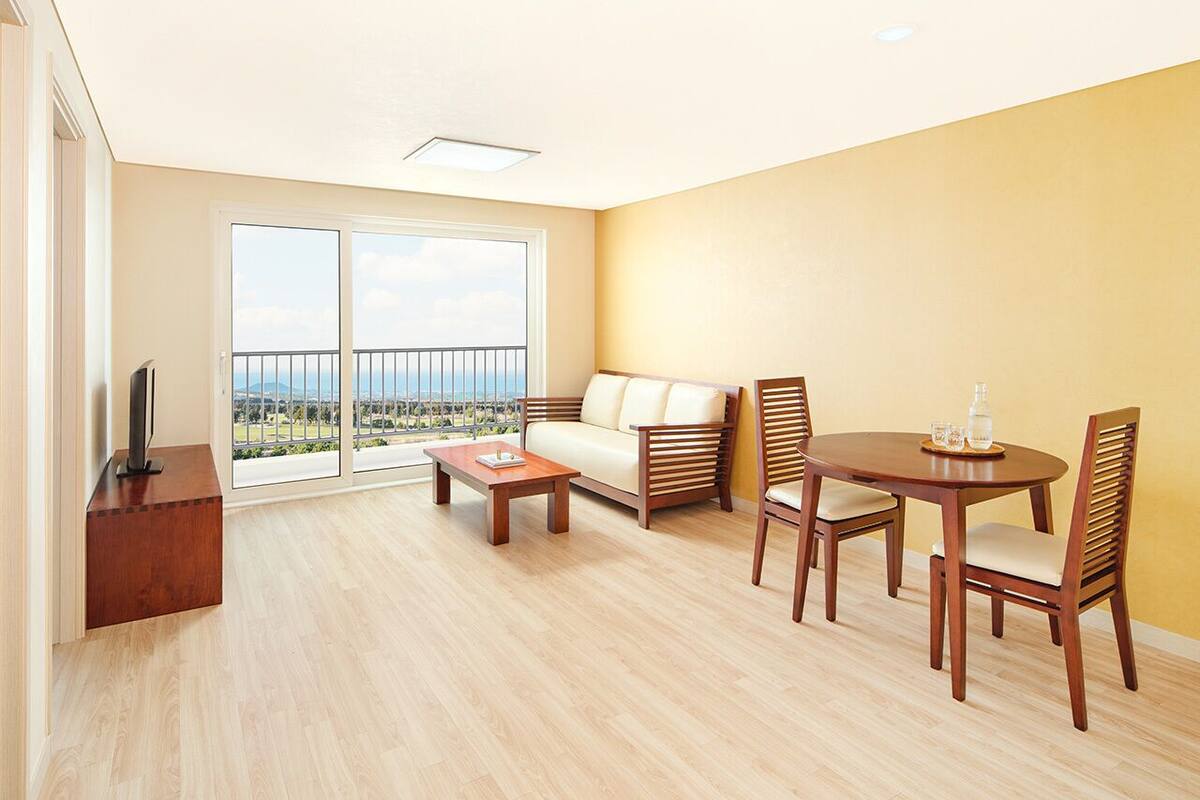 A spacious living room with a sofa, coffee table, and dining table with chairs. A flat-screen TV is on a wooden stand.
