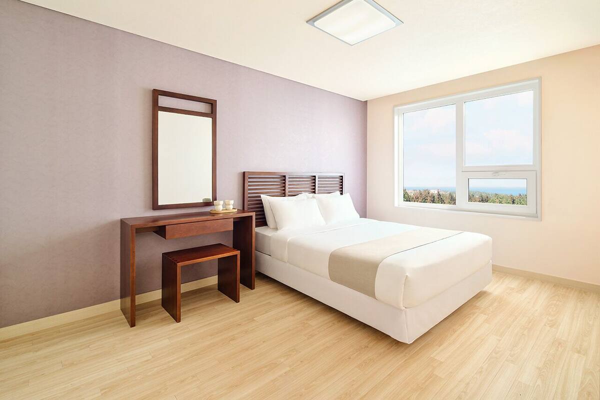 A bedroom with a bed, a desk, a chair, a mirror, and a window with a view.