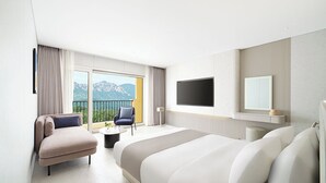 A modern hotel room with a large bed, a seating area with a chair and a small table, a flat-screen TV mounted on the wall, and a balcony with a view of mountains. - Hanwha Resort Seorak Sorano (Sokcho)