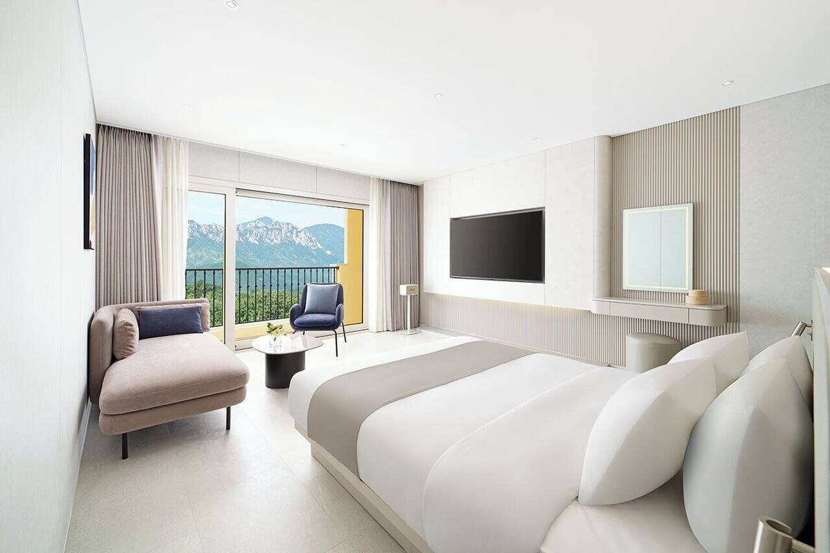 Ein modernes Hotelzimmer mit einem großen Bett, einer Sitzecke mit einem Sessel und einem kleinen Tisch, einem flachbildfernseher an der Wand und einem Balkon mit Bergblick.