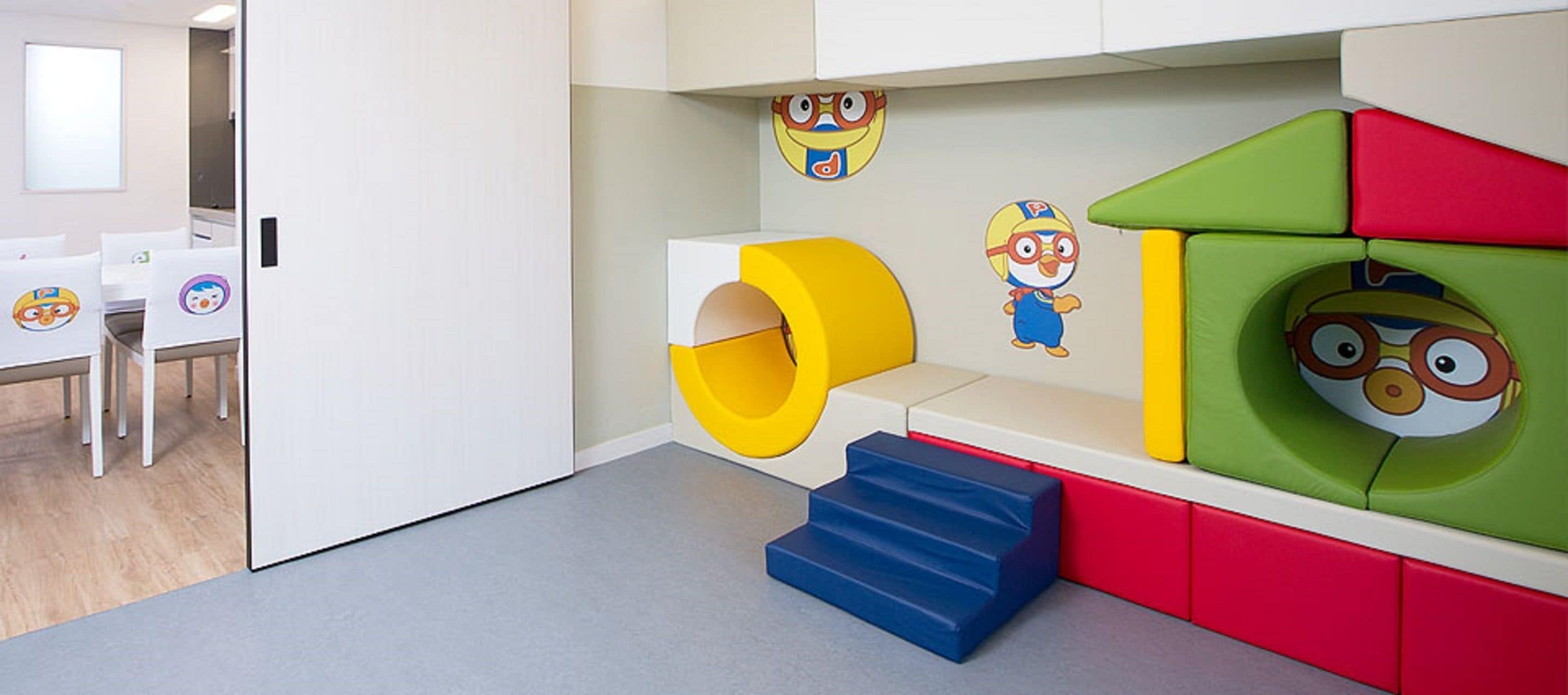 A children's play area with colorful play structures, including a slide and steps, and a wall decorated with cartoon characters.