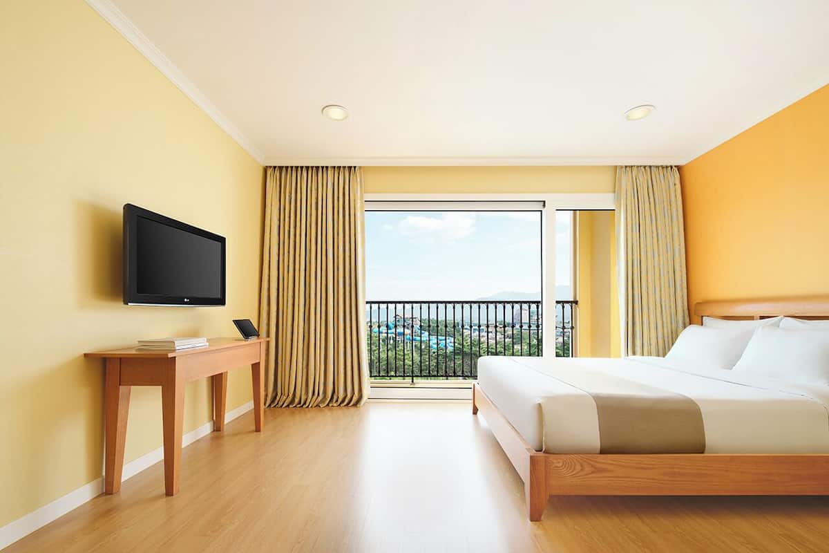 Ein Hotelzimmer mit Bett, Fernseher auf einem Holztisch, einem Balkon mit Vorhängen und Blick ins Grüne.