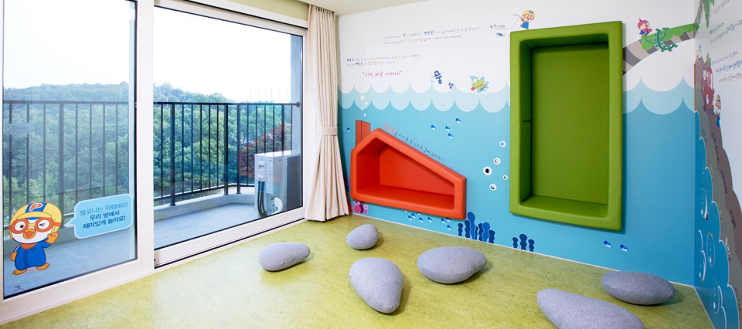 Zona de juegos infantiles con un ventanal grande, una puerta verde y vistas a un bosque.